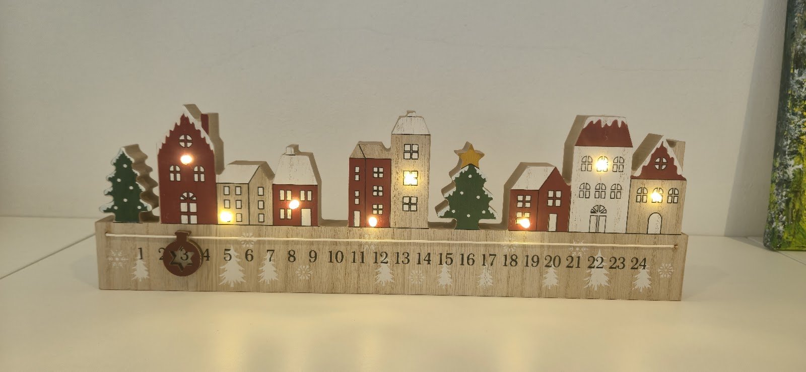 Dřevěná adventní dekorace v podobě svítícího městečka s očíslovanými domky na bílém podkladu.